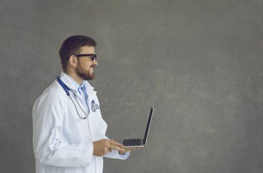 Portrait of confident smiling doctor physician in glasses white coat with stethoscope working using laptop side view shot on grey wall copy space. Online consultation service, telemedicine, e-health