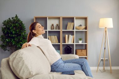 Happy woman sitting on a comfortable sofa at home. Side view of a young lady enjoying quiet leisure time and relaxing on a soft comfy couch in her beautiful living room