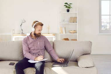 Focused young Caucasian man in headphones sit on sofa study online on laptop make notes. Guy in earphones work distant on computer at home office. Freelance job, remote education concept.
