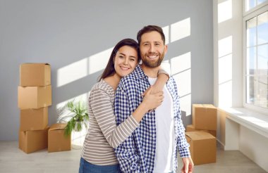 Portrait of a happy family couple in the new house on moving day. Young man and woman standing in a room with cardboard boxes in the background, looking at the camera and smiling. Real estate concept