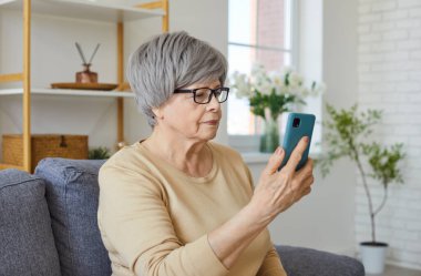 Serious elderly woman engages in digital leisure using phone for online communication or internet entertainment at home, focusing on connection and thoughtful approach to technology.