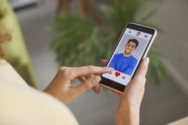 Single woman looking for man on online dating app. Young girl holding mobile phone, looking at profile pictures of male candidates and pressing heart buttons under photos she likes. Crop shot close up