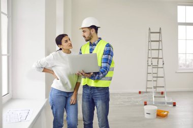 Attractive young woman is talking to construction foreman in white helmet, who demonstrates sequence of repairs in living room on laptop. Meeting of landlady of apartment with engineer or architect.