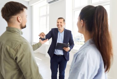 Smiling real estate agent hands keys to new home to couple, close-up. Couple rented apartment from realtor. Concept holding house, estate broker, apartment renters, giving keys, successful deal
