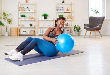Positive smiling young African American woman doing intense workout on yoga mat in living room at home, performing russian twist exercise with fitball for weight loss, core and abdominal muscles.