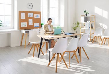 Businesswoman working online with a laptop, sitting at desk in an office. Her professional tasks involve financial inspection, internet research, and business operations in a focused environment.