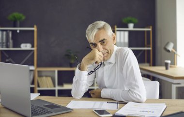 Portrait of a successful satisfied senior businessman sitting at his workplace in the office. Experienced manager who has successfully completed a working day sits smiling and looking at the camera.