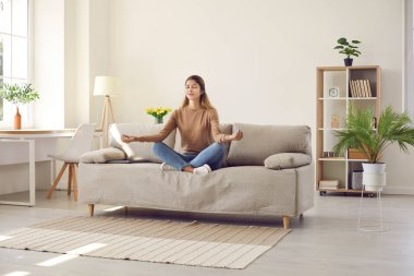 Calm young Caucasian woman sit on sofa in living room meditate practice yoga. Relaxed millennial girl in lotus position mudra hands breathe fresh air have meditation session. Stress free concept.