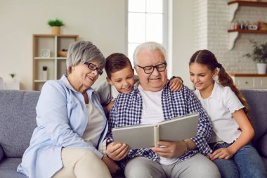 Happy family reading a book at home. Grandparents and children reading stories together. Smiling grandfather, grandmother and little kids sitting on the couch and looking at the book together