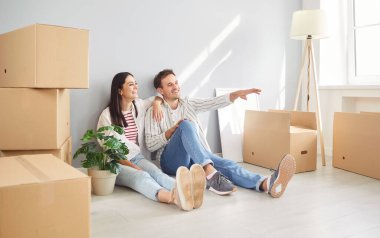 Happy young family sitting on empty floor among cardboard boxes in new house, planning, dreaming about buying interior design decorations, smiling couple enjoying moving in new apartment, relocation