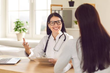 Portrait of smiling doctor sitting in office wearing stethoscope and holding report file with appointment and giving consultation to a woman sitting back during medical examination in clinic.