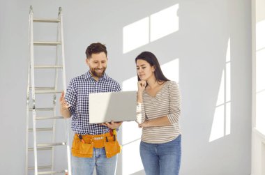 Genç evli çift taşınma gününden sonra boş bir odada bir apartman tadilatı ve tasarım projesi planlıyor. Gülümseyen mutlu adam ve kadın dizüstü bilgisayarla yeni evlerinin onarımını tartışıyorlar.