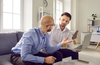 Young business men working on a laptop sitting on sofa in office. Two male employers talking and discussing new corporate projects or startups, analyzing company. Team work concept.