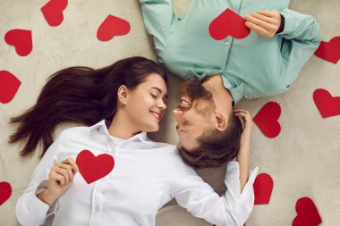 Happy beautiful couple spending St Valentines Day together. Young man and woman lying on floor among red heart shaped cards, looking at each other and smiling. Top view, high angle shot, from above