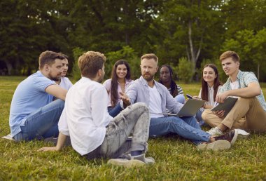 Group of students friends preparing for classes in nature or discussing social media in summer park. Diverse people sitting on the green grass outdoor with laptop together and talking.