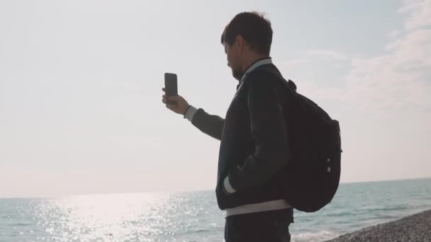 Um jovem fotógrafo tira fotos em um smartphone, um cavalheiro está no mar — Vídeo de Stock