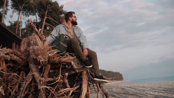 Hombre barbudo joven está sentado en ramas de madera en la playa de arena — Vídeo de stock