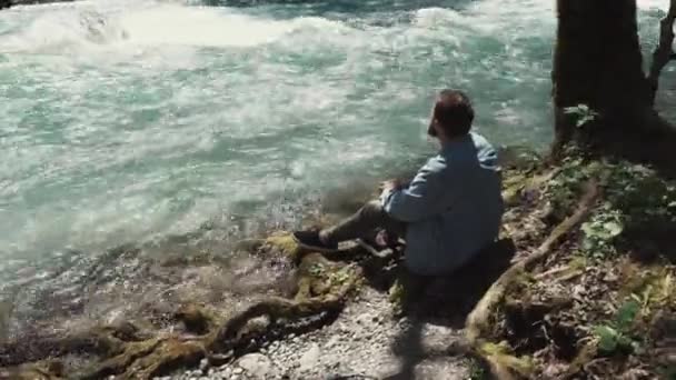 Молодий чоловік сидить біля швидкого потоку річки і дивиться на протікаючу воду — стокове відео