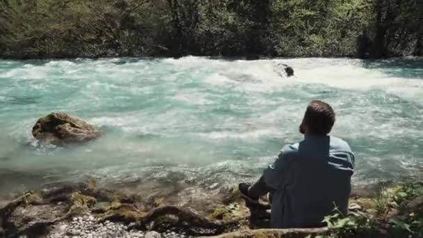 Один бородатый мужчина сидит на берегу и задумчиво смотрит на реку днем — стоковое видео
