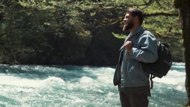 Un hombre que parece un guardabosques se para cerca de un río rápido y disfruta de un día de verano — Vídeo de stock