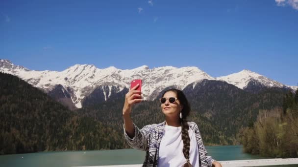 Joven bonita mujer está tomando selfies por su teléfono inteligente en el fondo de la montaña — Vídeos de Stock
