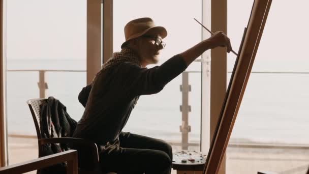 Artista profesional se dedica a dibujar sentado en una silla junto al mar — Vídeos de Stock