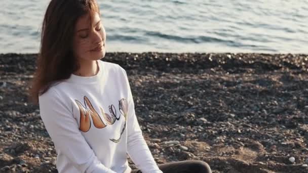Симпатичная и симпатичная женщина, сидящая на скальном пляже в свитере — стоковое видео