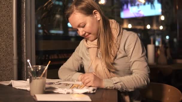 Blonde girl is painting on a white t-shirt by brush, sitting in cafe in evening — Stock Video