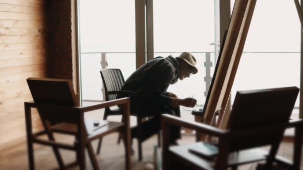Middelste leeftijd kunstenaar is het schilderen van een foto, zitten in zijn atelier — Stockvideo
