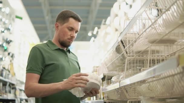 El hombre barbudo está eligiendo la lámpara en las tiendas de materiales de construcción, desempacándola — Vídeos de Stock