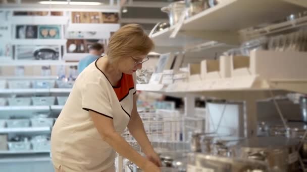 Alte Dame mit Brille inspiziert Metallpfanne in einem Geschäft mit Geschirr — Stockvideo