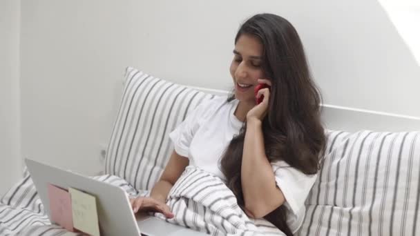 Menina está falando por telefone celular, sentado na cama de manhã, segurando laptop — Vídeo de Stock