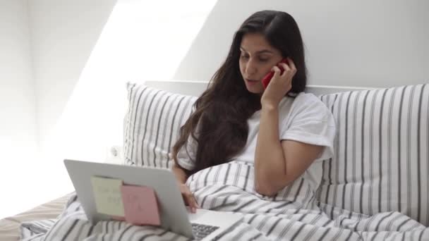 Веселая женщина работает с ноутбуком и мобильным телефоном, лежа в постели — стоковое видео