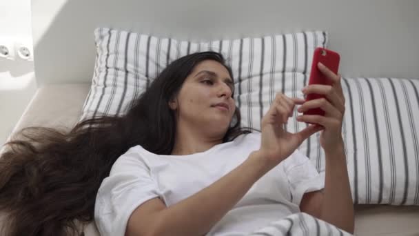 覚醒の若い女性はメッセージを入力して、スマート フォンでベッドで横になっています。 — ストック動画