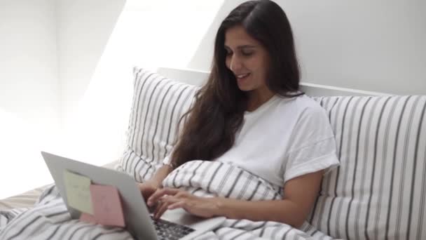 Lachende brunette zit in bed leunend op kussens en typen op laptop — Stockvideo