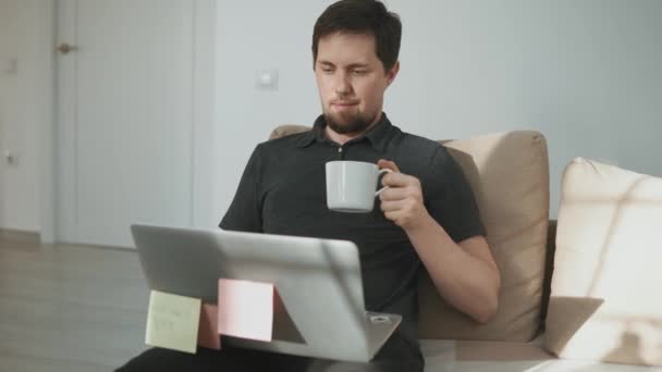 Молодой человек просматривает интернет на ноутбуке, сидя в гостиной с чашкой — стоковое видео
