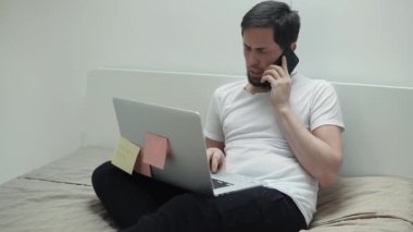 Meşgul bir adam cep telefonuyla konuşurken ve laptop, onun yatak odasında oturup yazmaya