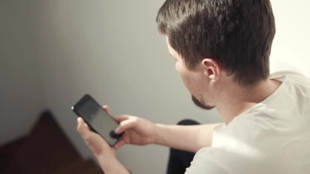 Молодой человек смотрит фотографии в своем смартфоне, сидит дома, смотрит сзади — стоковое видео