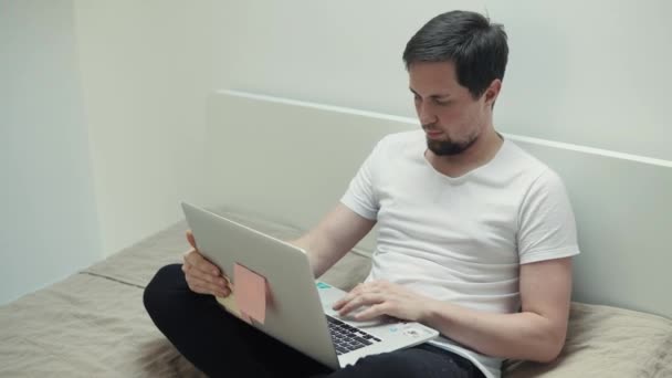 Μελαχροινός άντρας κάθεται στο κρεβάτι και εργάζεται με φορητό υπολογιστή στο σπίτι το βράδυ — Αρχείο Βίντεο