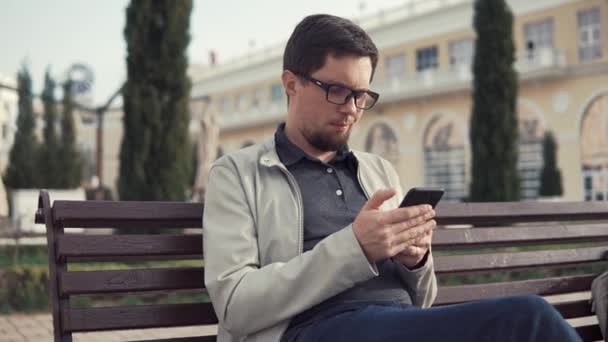 Hombre Brunet está leyendo mensajes y contestando con su teléfono inteligente, sentado en el parque — Vídeo de stock