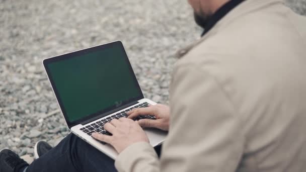 Man is sitting on gravel beach and working with notebook with chroma key screen — Stock Video