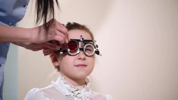 Oculista está poniendo vidrio verde y rojo en un marco de prueba en la cara de una niña pequeña — Vídeo de stock