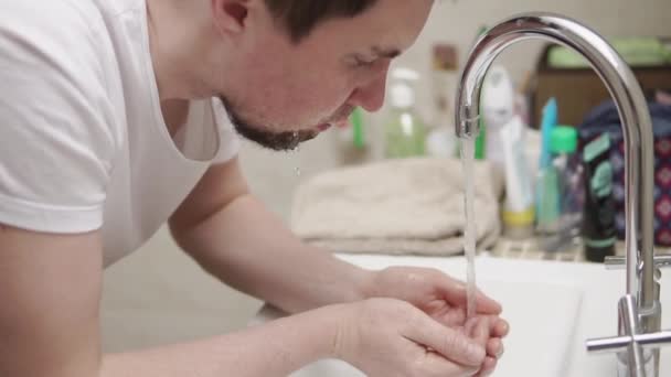 Een jonge en bebaarde man wast zijn gezicht onder de kraan in de badkamer — Stockvideo