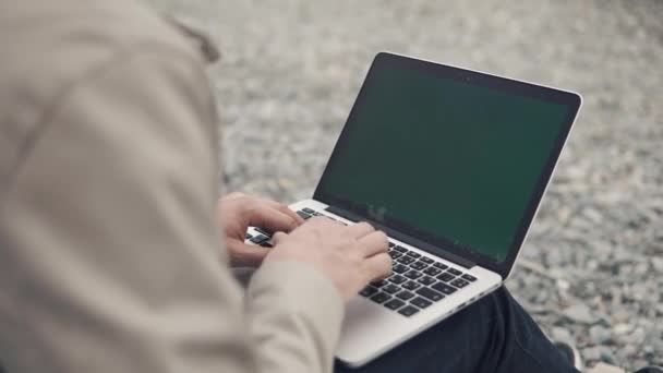 Nahaufnahme eines Mannes, der an einem Laptop im Freien zwischen einem felsigen Strand arbeitet — Stockvideo