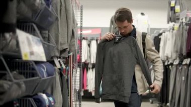 Erkek müşteri askı sportif kazak ile bir mağazada tutuyor. O kalite ve stil, büyük alışveriş merkezinde ticaret bir salonda ayakta inceliyor