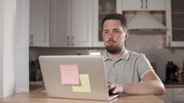 Взрослый веселый мужчина работает с ноутбуком в доме, сидит на кухне — стоковое видео