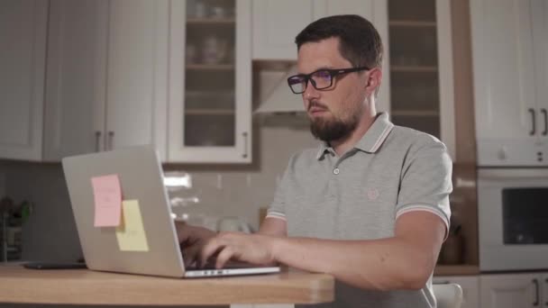 El hombre cansado está escribiendo en un portátil, sentado en casa, sacando sus gafas — Vídeo de stock