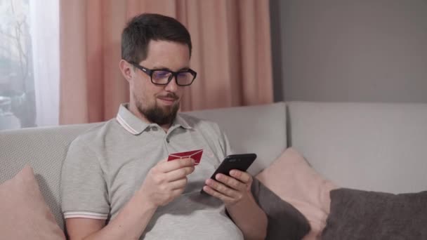 Adult man is sitting on couch in home and putting data of card in smartphone app — Stock Video