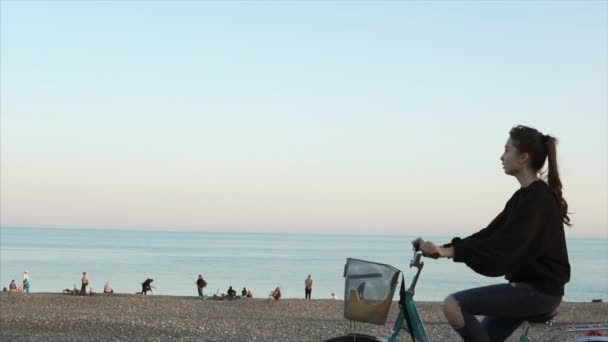 Adolescente chica está montando una bicicleta a lo largo de la playa de grava en la noche cerca del mar, vista lateral — Vídeos de Stock