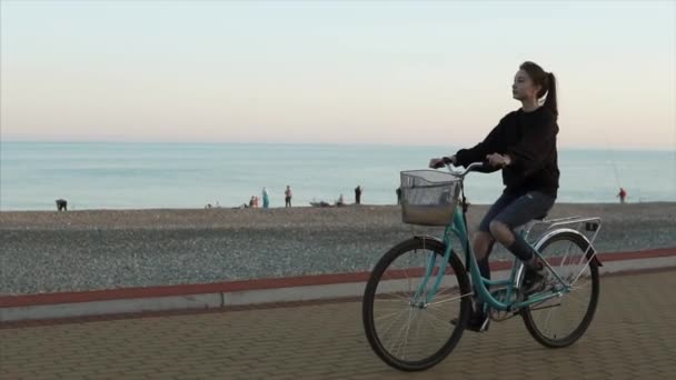 Tienermeisje is het berijden van een fiets langs kiezelstrand in avond in de buurt van de zee, zijaanzicht — Stockvideo